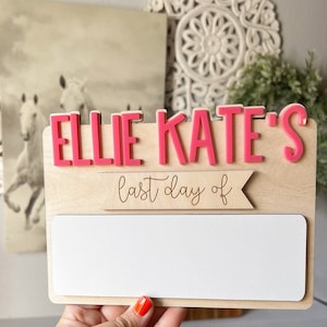 May include: A wooden sign with the name "Ellie Kate's" in pink letters. The sign also has a banner that says "last day of" and a blank white space for writing.