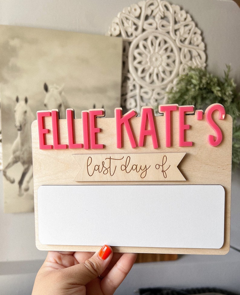 May include: A wooden sign with the name "Ellie Kate's" in pink letters. The sign also has a banner that says "last day of" and a blank white space for writing.
