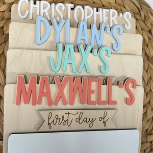 May include: Wooden sign with the names Christopher's, Dylan's, Jax's, and Maxwell's in different colors. The sign also has the text "first day of" and a blank space for writing.