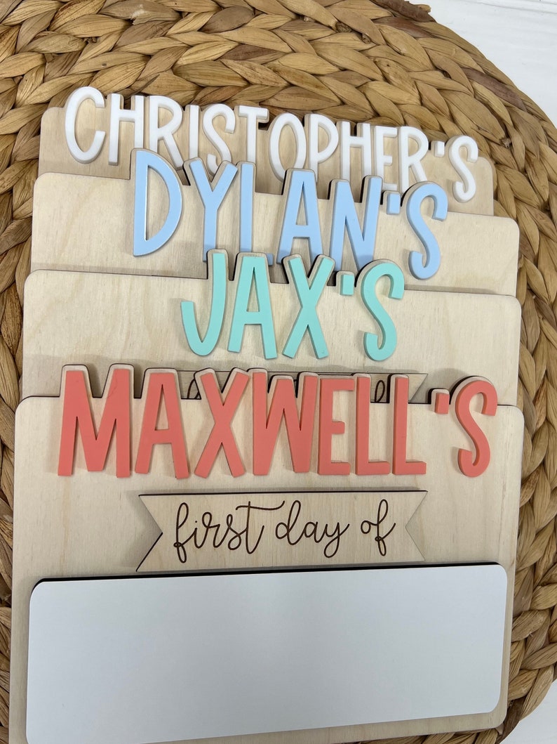 May include: Wooden sign with the names Christopher's, Dylan's, Jax's, and Maxwell's in different colors. The sign also has the text "first day of" and a blank space for writing.