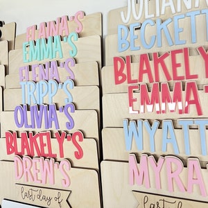 May include: A wooden board with a white dry erase surface and a banner that says "last day of". The board is decorated with colorful names, including "Juliana", "Beckett", "Blakeley", "Emma", "Wyatt", and "Myra".
