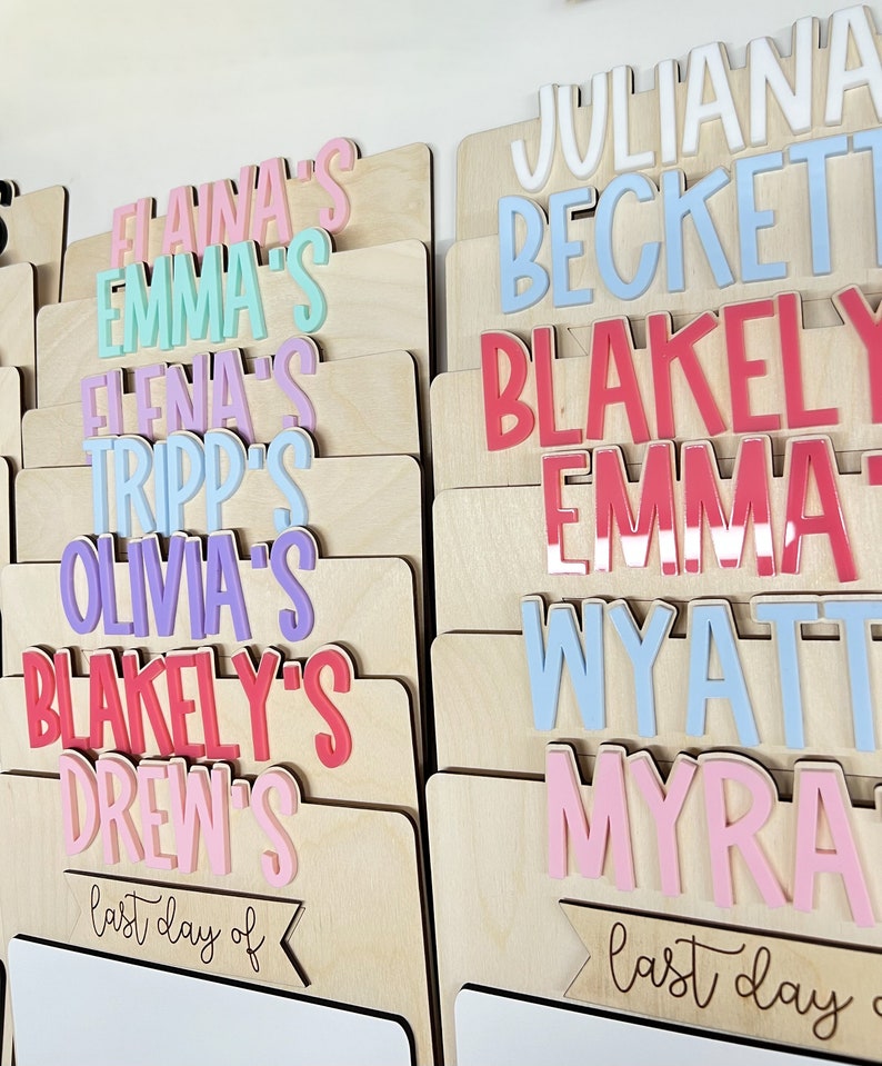 May include: A wooden board with a white dry erase surface and a banner that says "last day of". The board is decorated with colorful names, including "Juliana", "Beckett", "Blakeley", "Emma", "Wyatt", and "Myra".