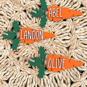 Puede incluir: Tres adornos de madera en forma de zanahoria con hojas verdes. Las zanahorias est&aacute;n pintadas de naranja y tienen letras blancas. Los nombres "Abel", "Landon" y "Olive" est&aacute;n escritos en las zanahorias.
