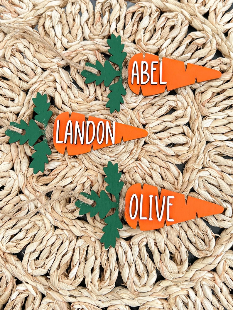Puede incluir: Tres adornos de madera en forma de zanahoria con hojas verdes. Las zanahorias est&aacute;n pintadas de naranja y tienen letras blancas. Los nombres "Abel", "Landon" y "Olive" est&aacute;n escritos en las zanahorias.