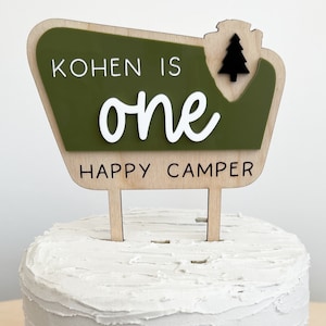 May include: A green and brown wooden cake topper with the text "Kohnen is one happy camper" and a small black pine tree.