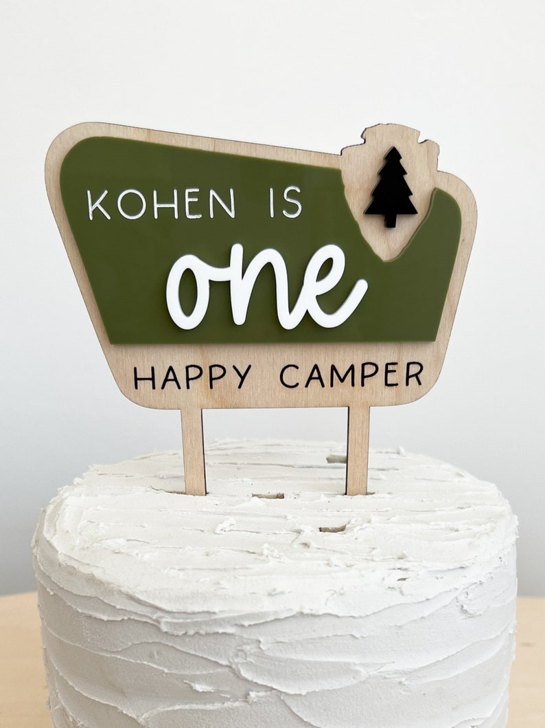 May include: A green and brown wooden cake topper with the text "Kohnen is one happy camper" and a small black pine tree.