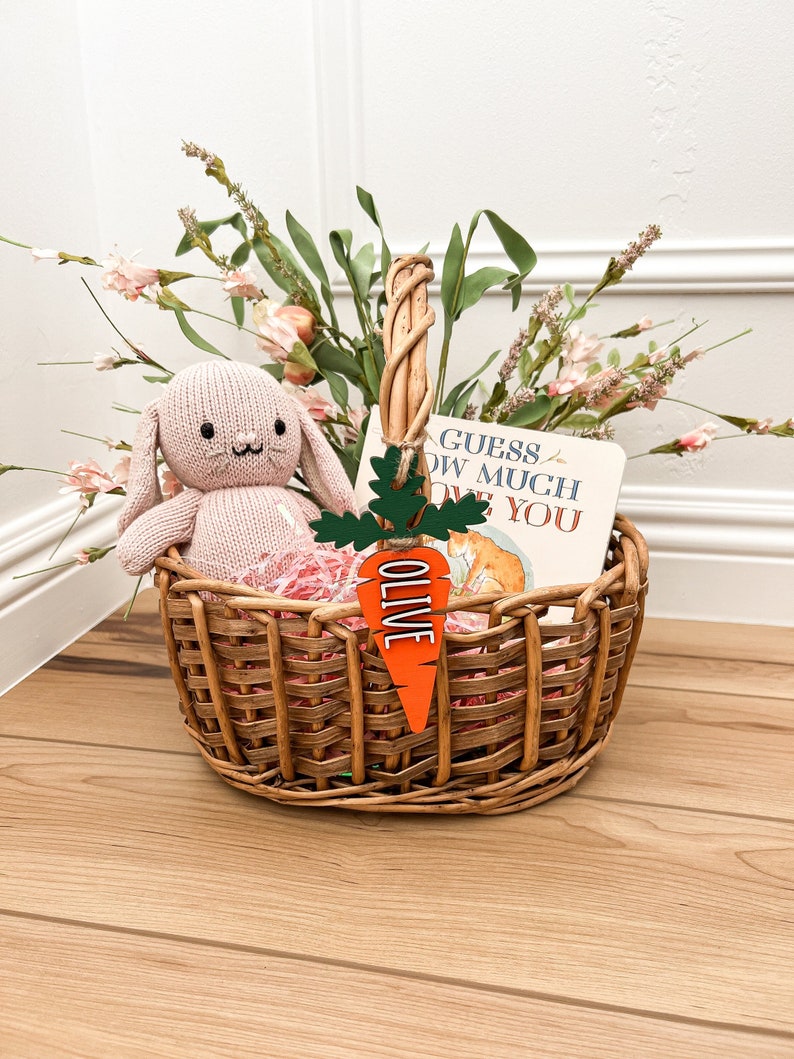 Puede incluir: Una cesta de mimbre llena de un conejito de peluche rosa, papel de seda rosa y un libro titulado "Adivina cu&aacute;nto te quiero". Una etiqueta de zanahoria de madera con el nombre "Olive" est&aacute; unida a la cesta.
