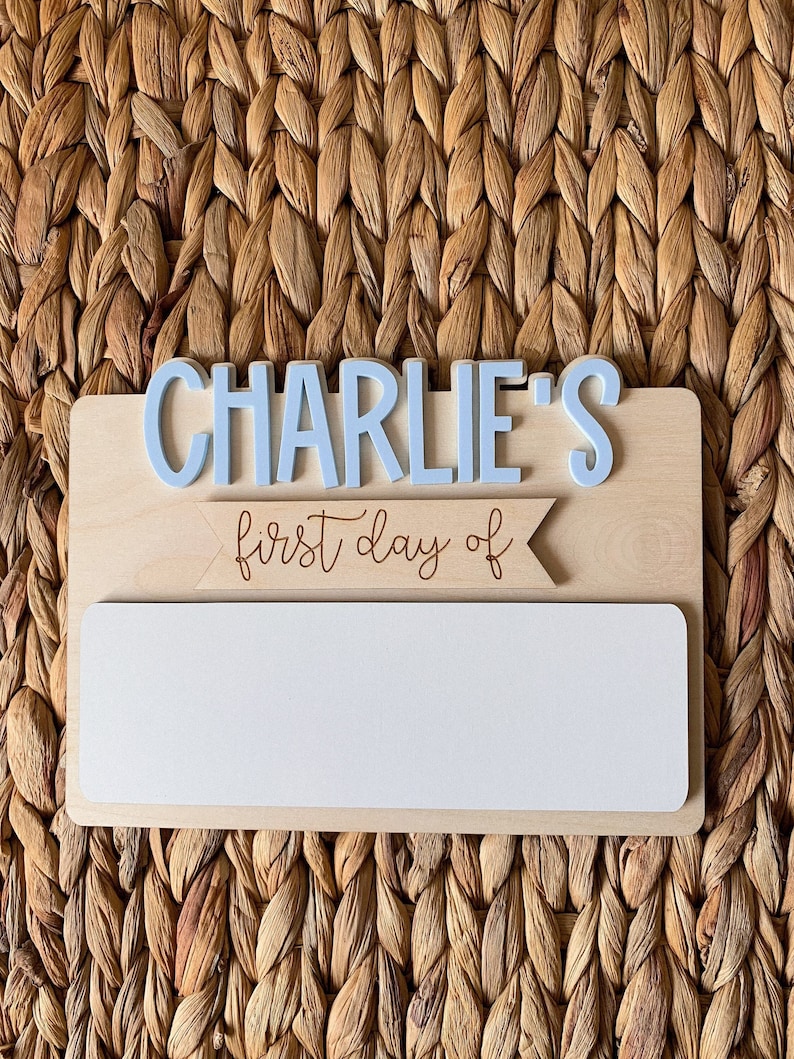 May include: A wooden sign with the text "CHARLIE'S first day of" in brown lettering. A blue banner with a white border is above the text. There is a blank white space below the text for writing.
