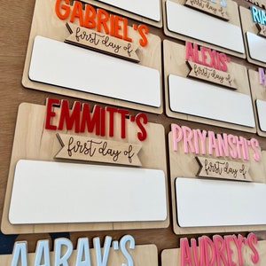 May include: A set of personalized wooden signs with different names and the text "first day of" in a decorative font. The signs are painted in various colors, including red, pink, orange, and white.