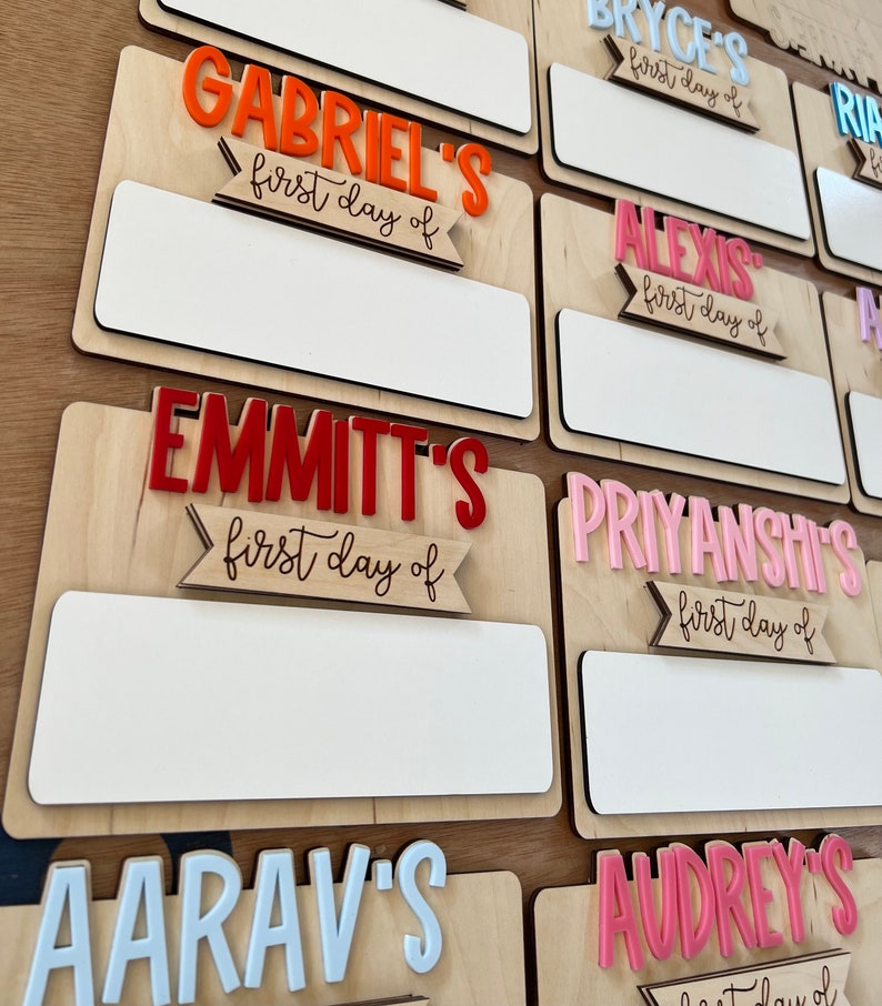 May include: A set of personalized wooden signs with different names and the text "first day of" in a decorative font. The signs are painted in various colors, including red, pink, orange, and white.