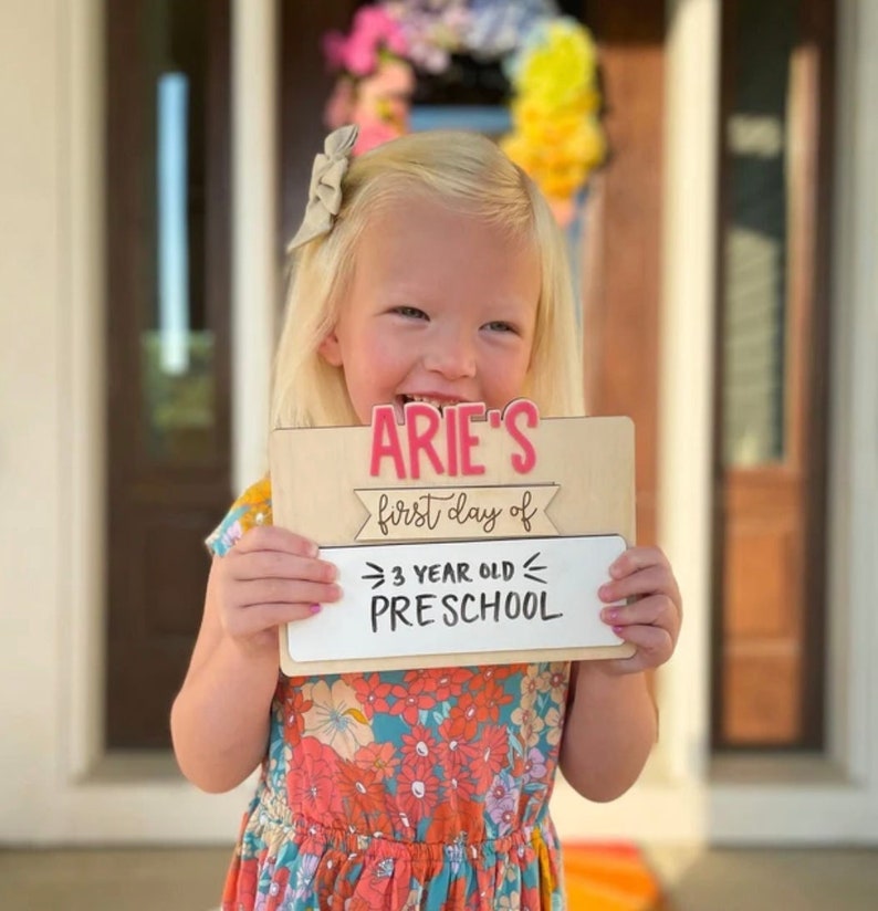 May include: A wooden sign with the text "Arie's first day of 3 year old preschool" in pink and black lettering. A young girl with blonde hair is holding the sign in front of her.