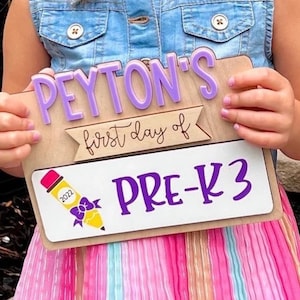 May include: A wooden sign with the text "PEYTON'S first day of PRE-K3" in purple and yellow lettering. The sign is decorated with a purple bow and a yellow pencil with the year 2022 on it.