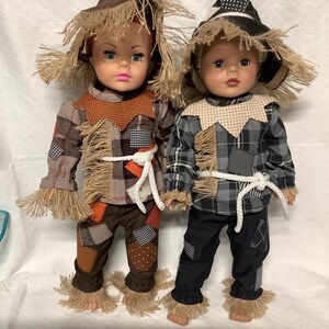 May include: Two dolls dressed as scarecrows. The left doll wears a brown plaid outfit with orange patches and a brown hat. The right doll wears a black and gray plaid outfit with a black hat. Both dolls feature straw-like fringe and white rope belts.