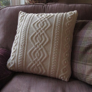 May include: A white knitted throw pillow with a cable knit pattern sits on a purple couch with a plaid throw pillow.