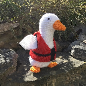 May include: A white knitted goose wearing a red life vest with black trim. The goose is standing on a rock wall.