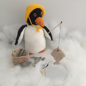 May include: A crocheted penguin wearing a yellow hat holds a fishing rod with a crocheted fish on the line. The penguin is surrounded by a white fluffy surface and a basket of crocheted fish.