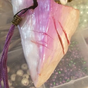 Puede incluir: Un colgante en forma de corazón iridiscente morado y blanco con un cordón morado y un pequeño dije de metal. El colgante está en una caja con varias cuentas.