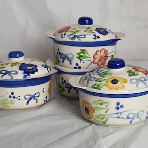 May include: A set of four ceramic casserole dishes with lids. The dishes are off-white with a blue rim and handles. Each dish is decorated with hand-painted flowers in red, yellow, and blue, along with blue bows and green leaves. The lids have a blue knob.