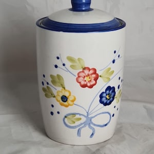 May include: White ceramic canister with a lid, decorated with a floral pattern. The flowers are red, yellow, and blue, with green leaves and blue accents. The lid and rim have a blue stripe.