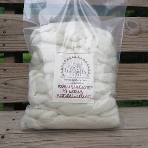 May include: A clear plastic bag filled with white merino wool top. A label on the front features the text Wet Belly Fibers, 100% U.S. Merino Top, 19 Microns, Natural, 1 Pound, and the website address wetbellyfibers.com with a logo of animals.