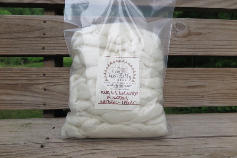 May include: A clear plastic bag filled with white merino wool top. A label on the front features the text Wet Belly Fibers, 100% U.S. Merino Top, 19 Microns, Natural, 1 Pound, and the website address wetbellyfibers.com with a logo of animals.