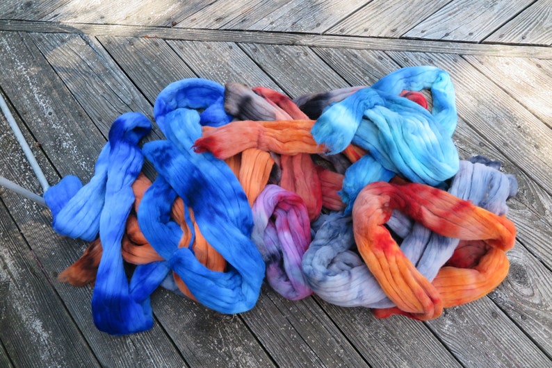 May include: A collection of hand-dyed wool roving fiber in vibrant shades of blue, orange, red, and purple. The soft, unspun textile material is arranged in loose, tangled loops, showcasing a gradient of saturated colors and blended tones.