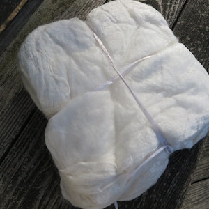 May include: A square-shaped bundle of white, fibrous material with a soft, textured surface. The bundle is secured with thin, white ribbon or string tied in a cross pattern across the top, creating four distinct, slightly puffed sections.