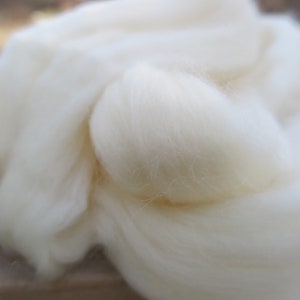 May include: A soft, fluffy mass of unspun white wool fiber. The material has a cloud-like, airy texture with fine, delicate strands that are loosely gathered together in a voluminous pile, showcasing the natural, raw state of the white fiber.