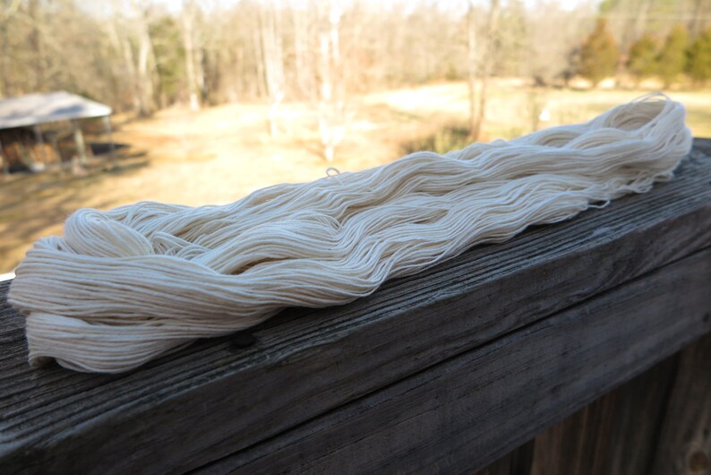 Peut inclure: Un long &eacute;cheveau continu de fil blanc cass&eacute; pr&eacute;sentant une texture douce et torsad&eacute;e. Le fil est compos&eacute; de multiples brins fins individuels regroup&eacute;s en une boucle l&acirc;che et allong&eacute;e, mettant en valeur une teinte naturelle, cr&egrave;me et neutre.