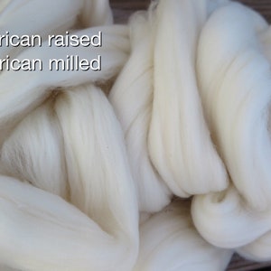 May include: A large, soft pile of unspun white wool roving with a fibrous, fluffy texture. The text "American raised American milled" is printed in white letters on the upper left side of the image, clearly visible against the white wool fibers.