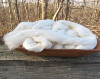 May include: A large, fluffy pile of white, unspun wool roving is contained within a smooth, elongated, dark brown wooden bowl. The wool has a soft, fibrous texture and is piled high, filling the entire interior space of the wooden vessel.