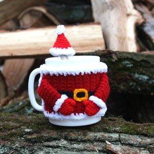 Può includere: Una tazza in ceramica bianca con un design a crochet di Babbo Natale. La tazza ha un vestito di Babbo Natale rosso e bianco con una cintura nera e una fibbia gialla. Babbo Natale ha una barba bianca e un cappello rosso.