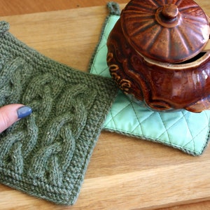 以下が含まれることがあります： ケーブル編みのパターンが施された緑色のニット製の鍋つかみが2つ。1つの鍋つかみは木製の表面に置かれ、もう1つは蓋付きの茶色の陶器の鍋の下にある。