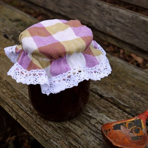 Peut inclure: Un pot de confiture foncée recouvert d'un couvercle en tissu à carreaux et d'une bordure en dentelle blanche. Une cuillère en bois avec un motif peint repose à côté du pot sur une surface en bois vieilli. Le pot semble fait maison.