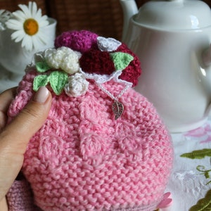 May include: A pink knitted tea cosy with a crocheted flower embellishment. The cosy is decorated with white, pink and green crocheted flowers and a silver charm.