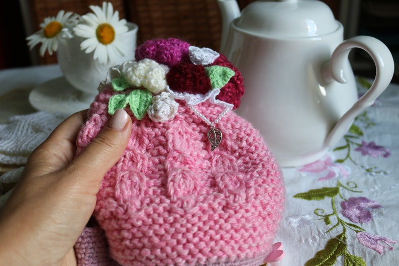 May include: A pink knitted tea cosy with a crocheted flower embellishment. The cosy is decorated with white, pink and green crocheted flowers and a silver charm.