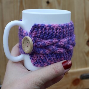 May include: White ceramic mug with a purple and pink knitted cozy. The cozy has a wooden button with the text "With Love" on it.