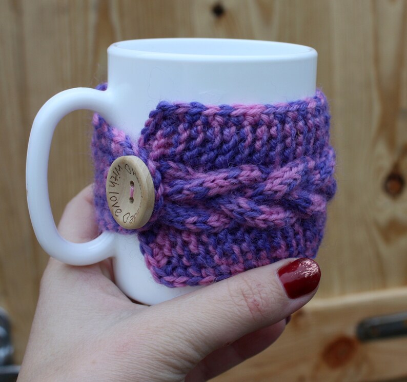 May include: White ceramic mug with a purple and pink knitted cozy. The cozy has a wooden button with the text "With Love" on it.