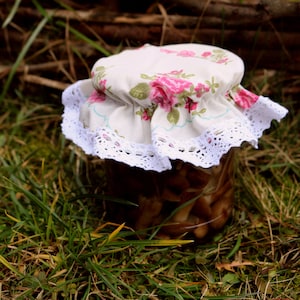 Puede incluir: Un tarro de vidrio lleno de champiñones, cubierto con una tapa de tela blanca con estampado floral rosa y ribete de encaje.