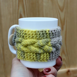 May include: A white ceramic mug with a knitted cozy in yellow and brown. The cozy has a cable knit pattern.