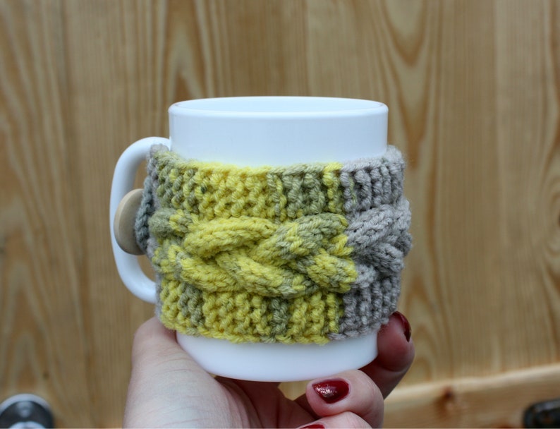 May include: A white ceramic mug with a knitted cozy in yellow and brown. The cozy has a cable knit pattern.