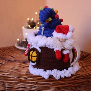 May include: Crocheted mug cozy in the shape of a house with a gnome wearing a red hat and a blue tree on top. The cozy has a brown base, a white snowy roof, and a yellow window.