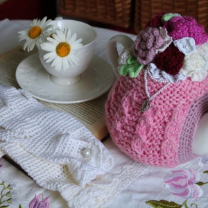 May include: A pink crocheted teapot cosy with floral embellishments, a white teapot, a white crocheted lace glove, and a book with daisies in a white teacup.