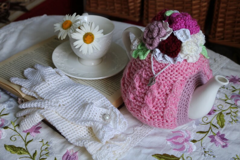 May include: A pink crocheted teapot cosy with floral embellishments, a white teapot, a white crocheted lace glove, and a book with daisies in a white teacup.