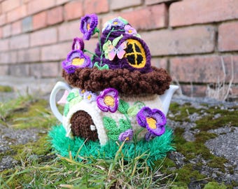 Puede incluir: Una encantadora funda de tetera hecha a mano, con un diseño de casa de hadas en miniatura. La funda está adornada con flores moradas, hojas verdes y un tejado de paja marrón. La tetera es blanca y la funda se asienta sobre un lecho de hilo verde.