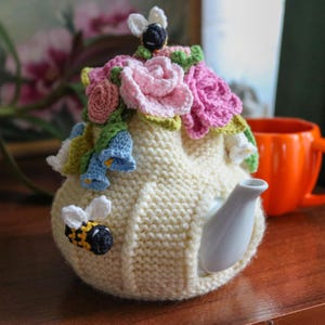 Puede incluir: Un calienta-tetera de crochet blanco con un diseño floral. El calienta-tetera presenta flores rosas, blancas y azules, así como pequeñas abejas de crochet.