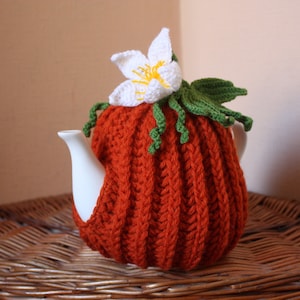 Puede incluir: Un calienta-tetera de punto de ganchillo en forma de calabaza con una flor blanca y hojas verdes en la parte superior. El calienta-tetera es de color naranja y está sobre una superficie de mimbre.