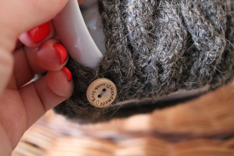 May include: A close-up of a brown knitted mug cozy with a wooden button that says "Handmade with love" in cursive.