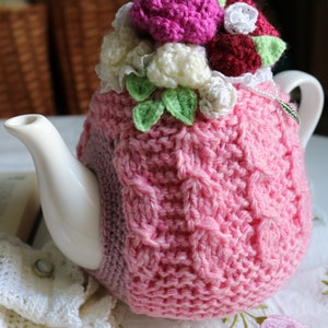 May include: A pink crocheted teapot cosy with a floral design. The cosy features white, pink and burgundy flowers with green leaves. The cosy is on a white teapot.