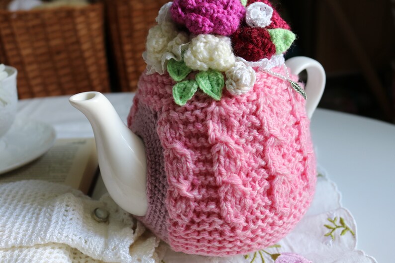 May include: A pink crocheted teapot cosy with a floral design. The cosy features white, pink and burgundy flowers with green leaves. The cosy is on a white teapot.